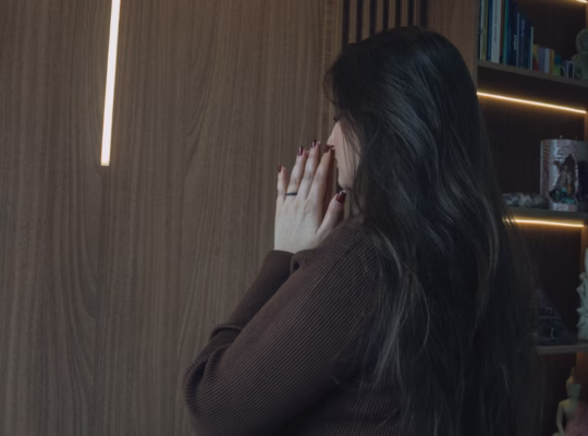 A woman prays in a serene room with an illuminated cross and open Bible.